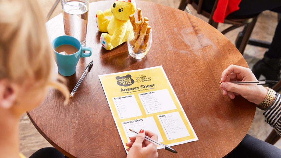 Two people filling in the answers on a Big BBC Quiz answer sheet on a table in a cafe.