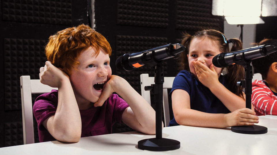 Two children laughing in front of microphones in a recording studio