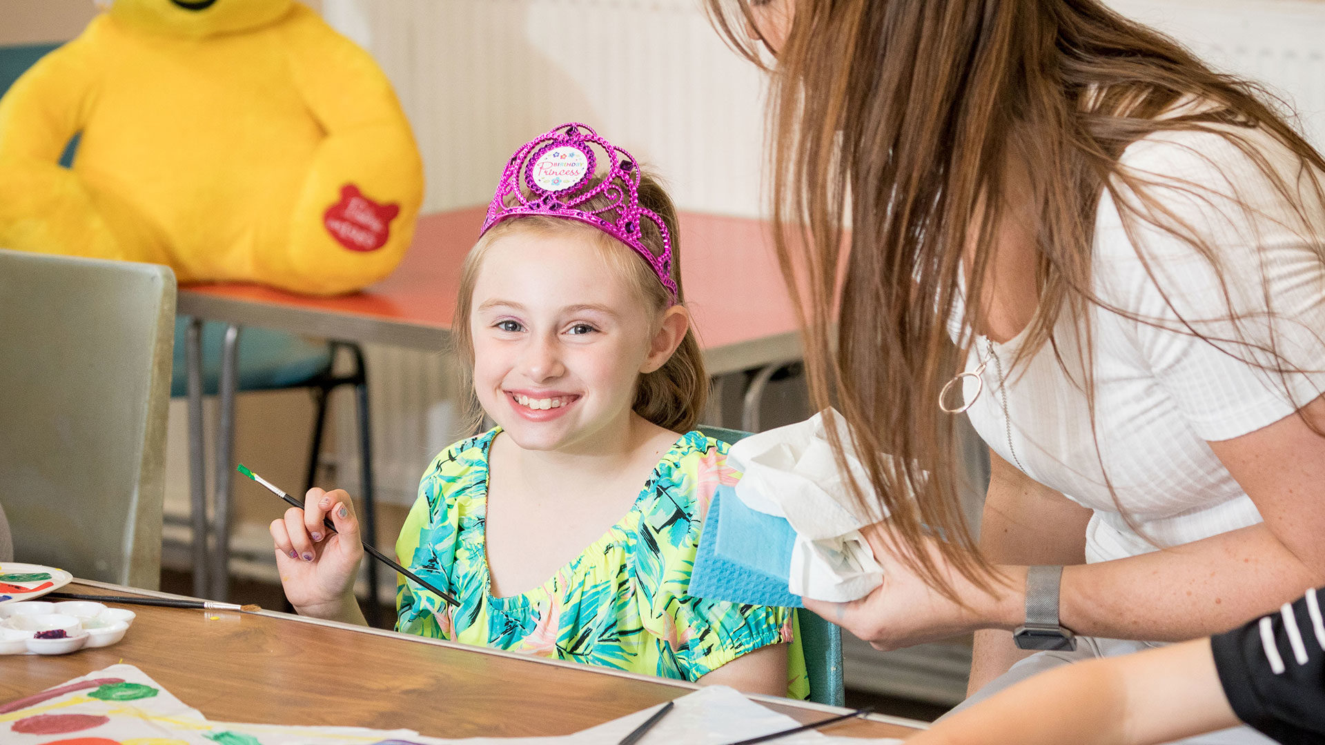 Young smiling girl dressed as a princess, painting with a female volunteer