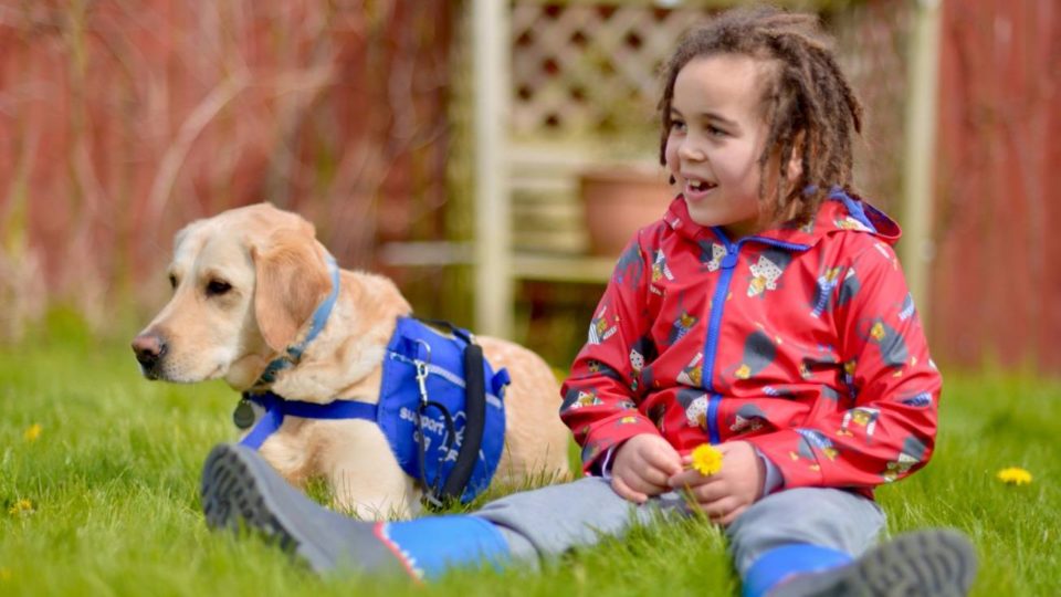 A young boy in a coat and wellies sat in the grass with his support dog