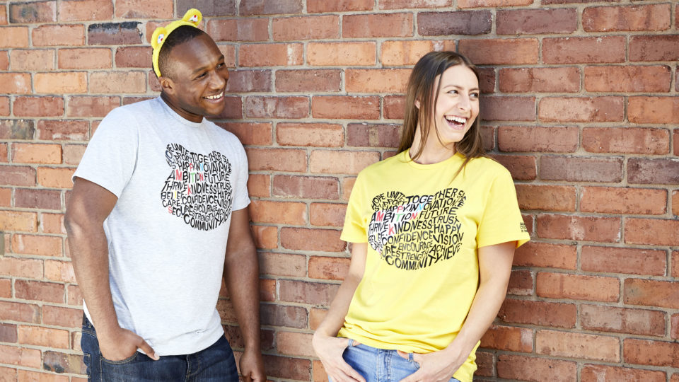 A man and a woman wearing the 2019 BBC Children in Need fundraising t-shirts and leaning against a brick wall