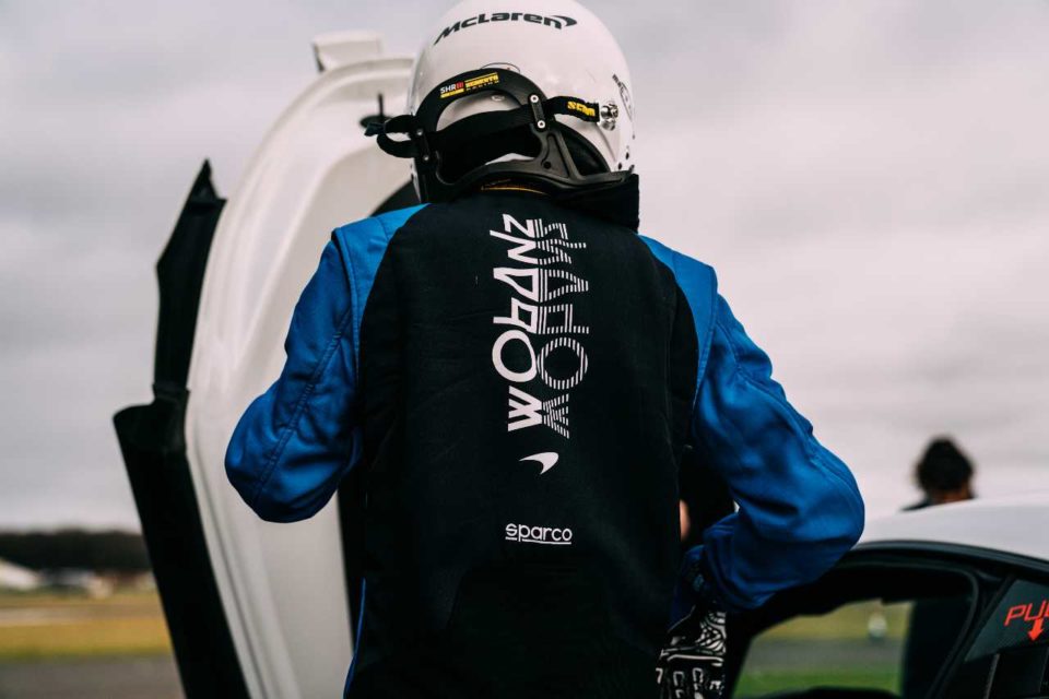 Someone dressed in a car racing suit wearing a helmet with the Shadow Project logo on the back of the suit