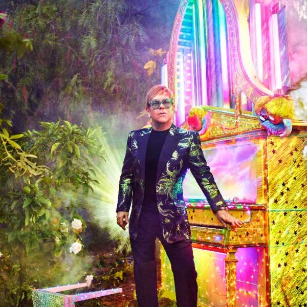 Elton John leaning against a piano surrounded by greenery and coloured lights