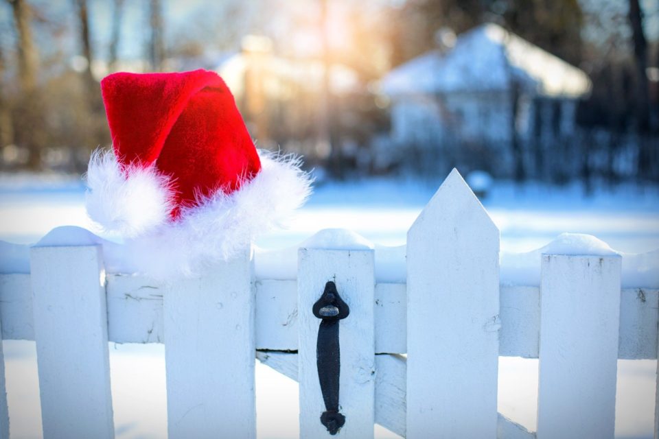 A Santa hat hung on a white gate with a snowy background and a house in the distance