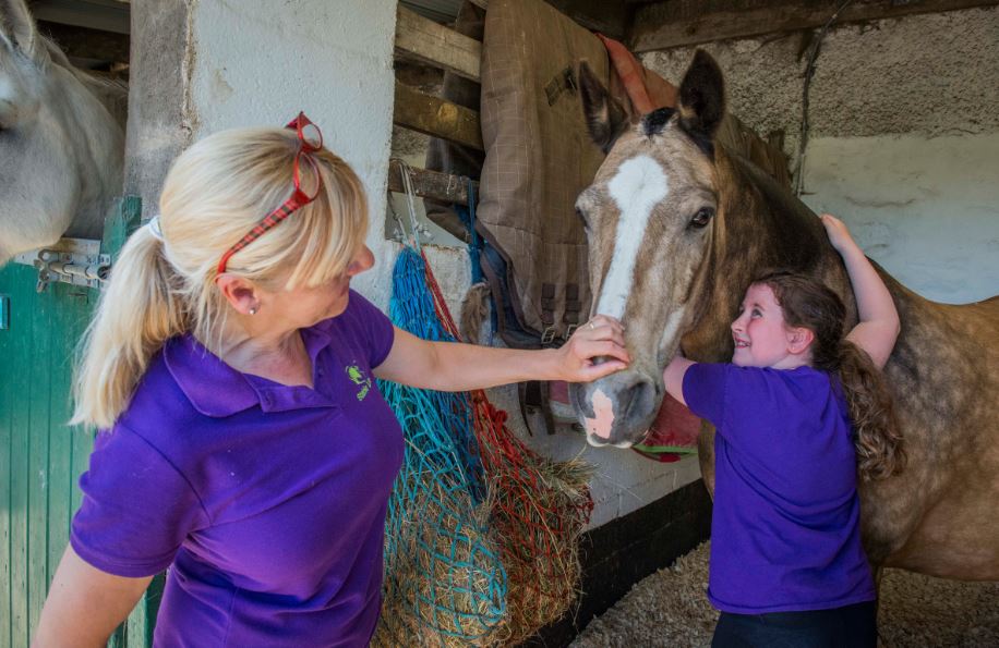 Young people from Stable Life learn how to look after a horse in a stable, one of them is lifting the saddle onto the horses back and another is being shown how to put on the bridle. They are all wearing purple tops.