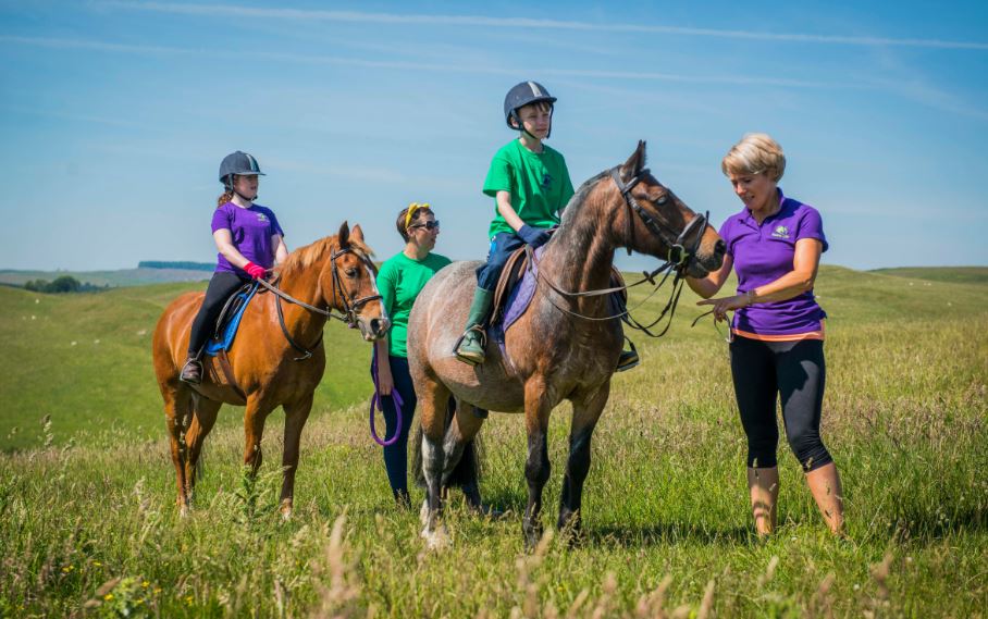 Young people ride horses out in a field. The sun is shining and the sky is bright blue. Project workers lead the horses through the field.