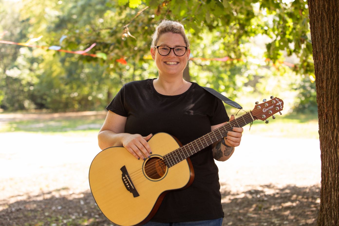 Emily stood with a guitar in a leafy park on a sunny day, wearing a black t-shirt, glasses and her blonde hair tied back