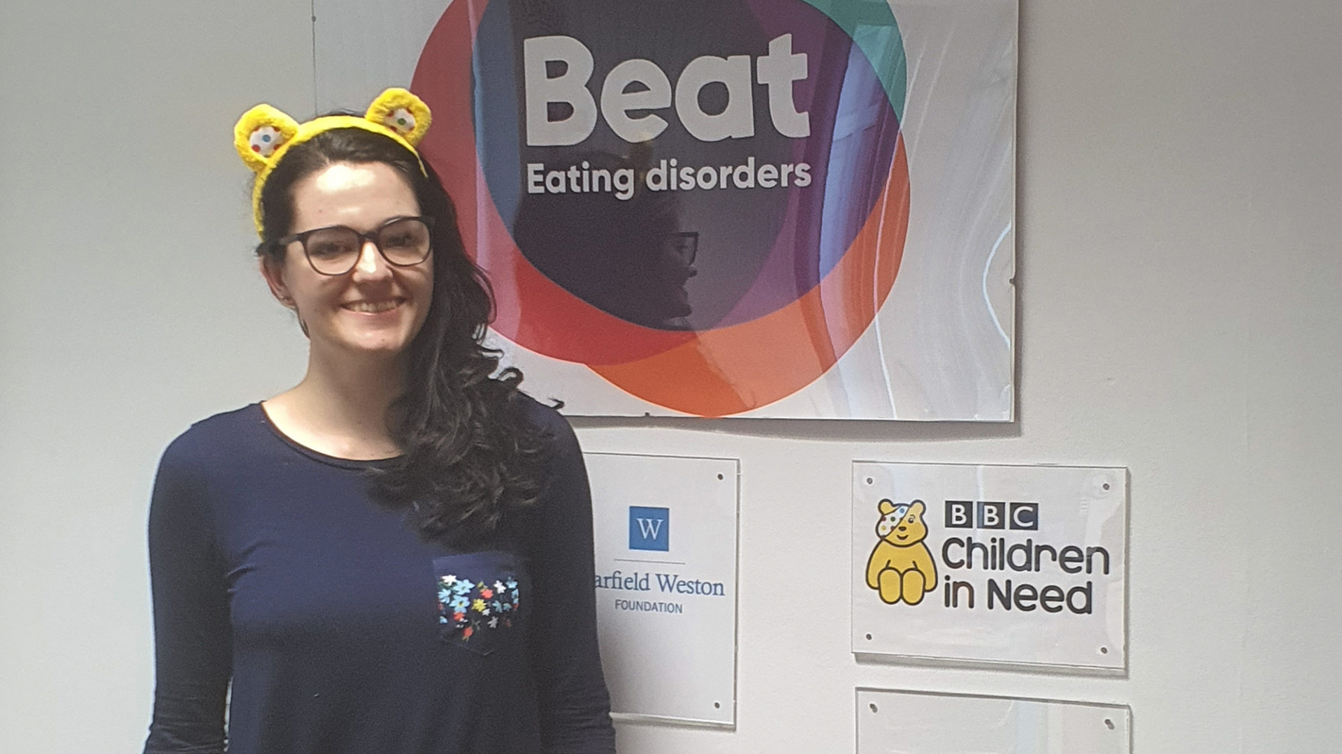 A photo of Tash wearing Pudsey ears and standing in front of the Beat sign