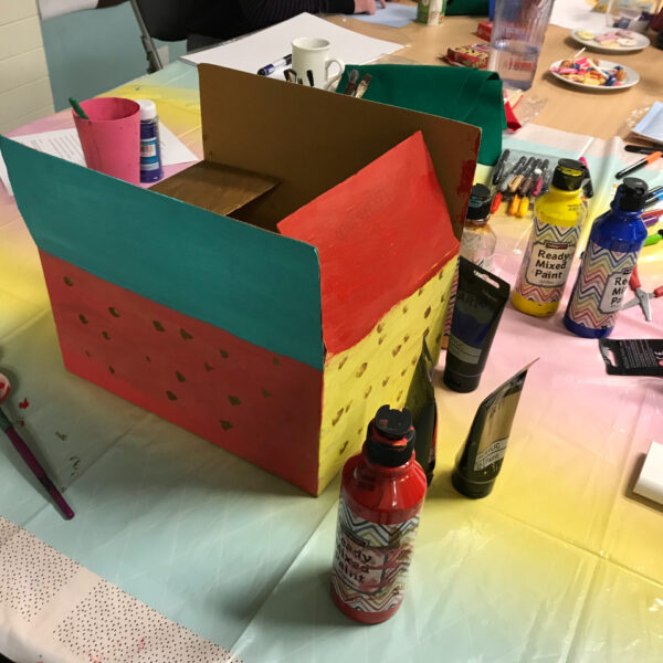 A cardboard box decorated with paint, surrounded by bottles of paint