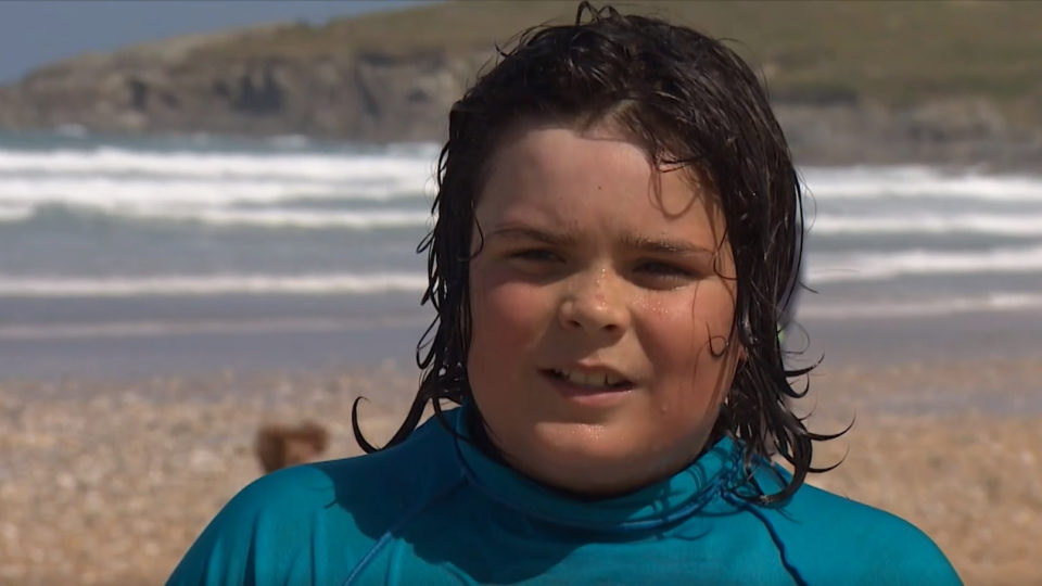 Skyler dressed in a wet suit with wet hair, standing on a beach