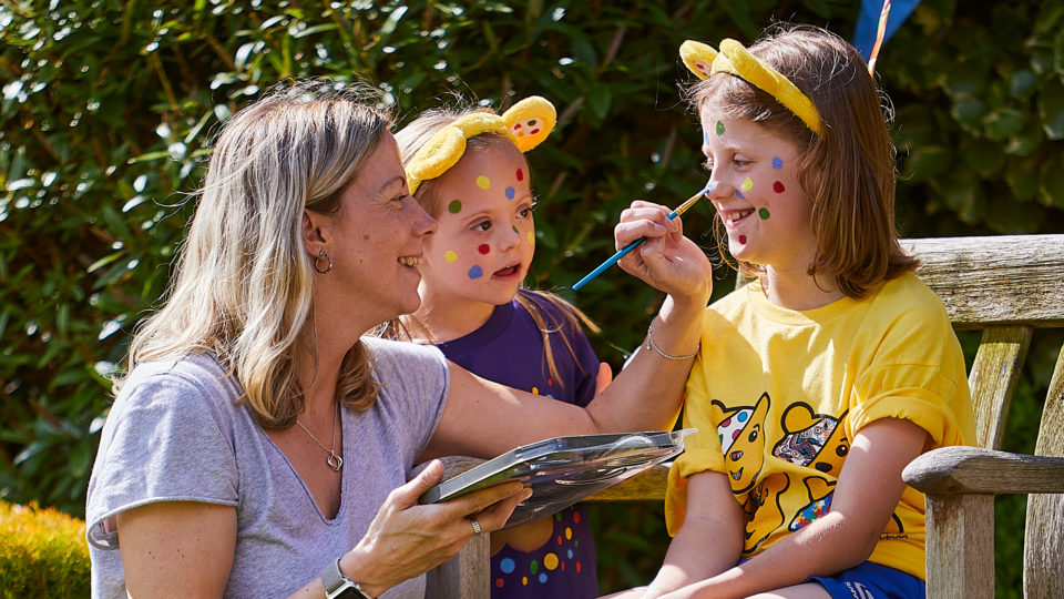 A lady paints coloured spots on the face of two girls wearing Pudsey ears