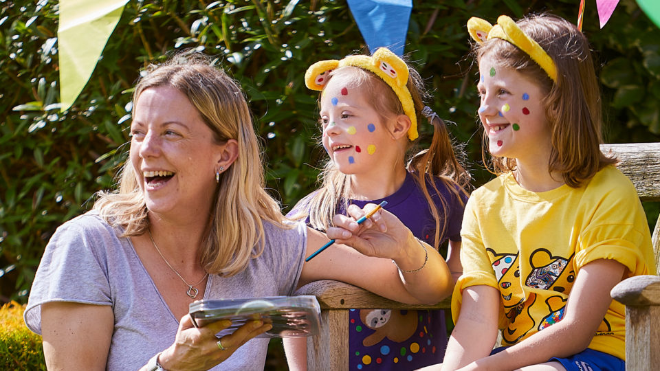 A lady paints coloured spots on the face of two girls wearing Pudsey ears