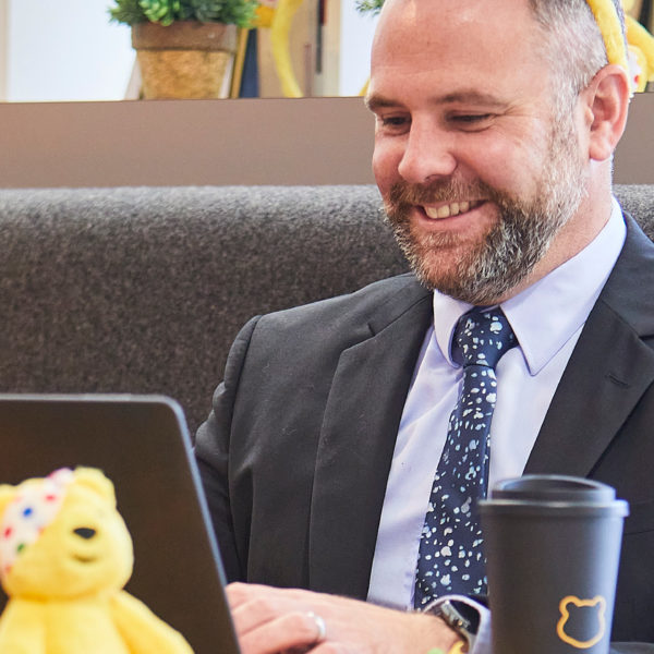 man in a suut on a laptop smiling with a Pudsey plush toy