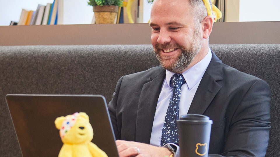 man in a suut on a laptop smiling with a Pudsey plush toy
