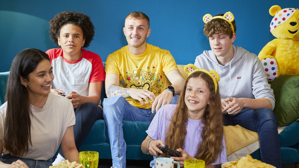 Young people sit on the floor or a sofa playing video games with yellow t-shirts, ears and a Pudsey Bear