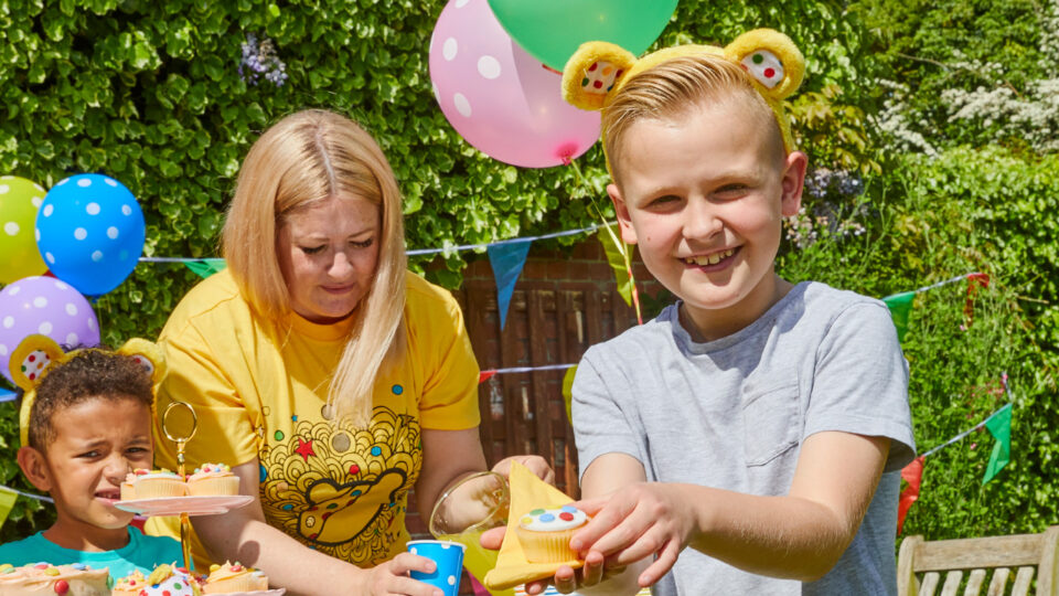 A young oy wearing yellow Pudsey ears holds up a spotty cupcake to the camera smiling