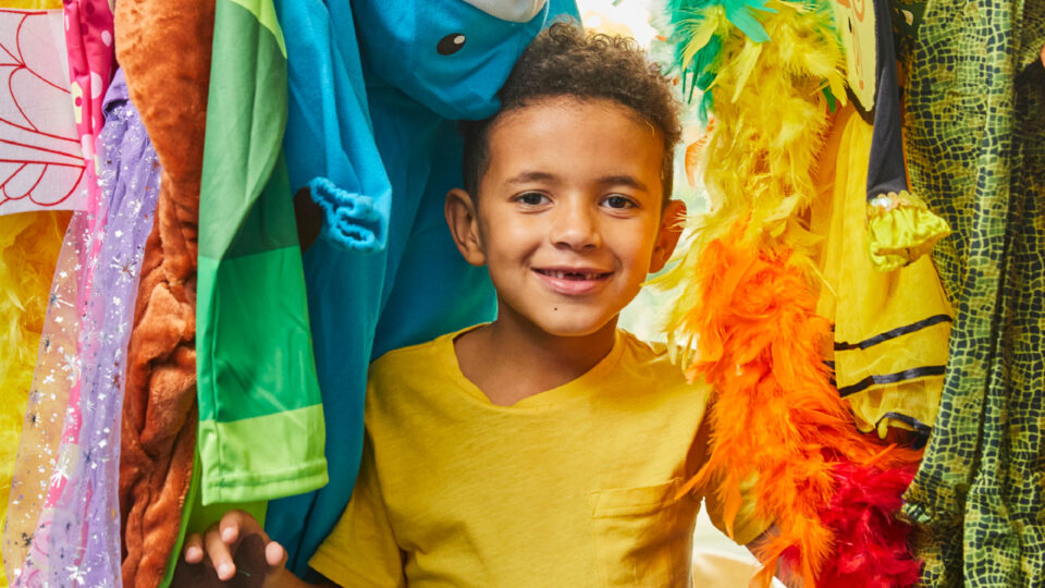 A young boy with two front teeth missing smiles as he pops out from a rack of fancy dress costumes