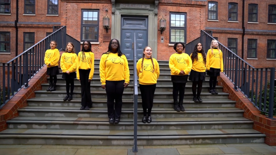 the Children in Need choir in yellow t-shirts on the steps of a building singing