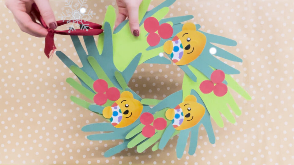 A screenshot from the craft video showing the Christmas wreath made from green paper hands arranged in a circle, with red paper berries and Pudsey bear faces as decoration