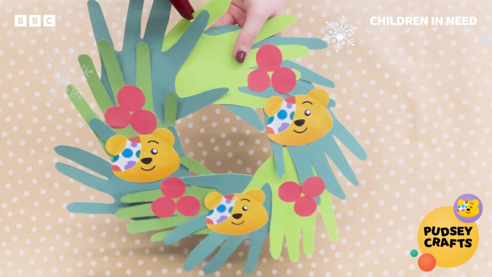 A screenshot from the craft video showing the Christmas wreath made from green paper hands arranged in a circle, with red paper berries and Pudsey bear faces as decoration