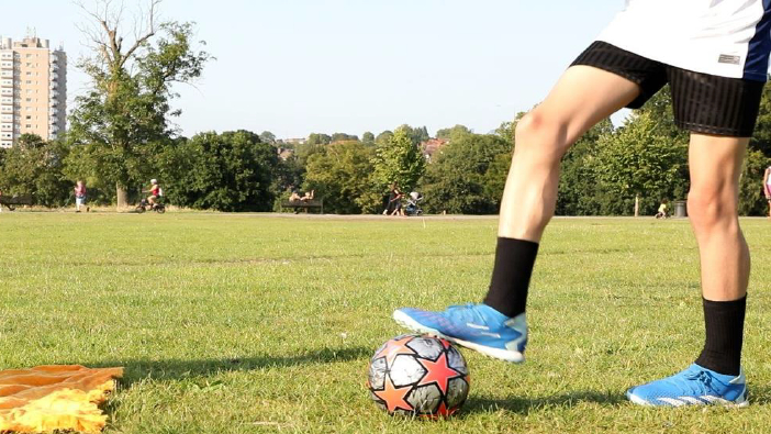 a young person with their foot on a football in a park with a high rise building in the background