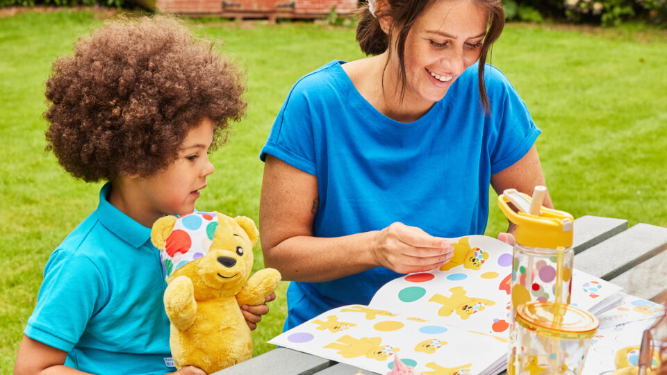 a child holding a pudsey bear looking at a Pudsey sticker book with a lady