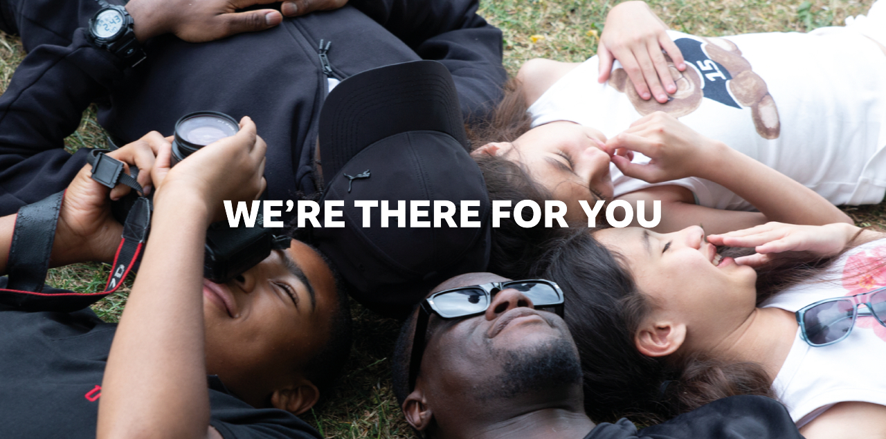Young people lying on the grass smiling. One holds a camera, one is wearing sunglasses. The words 'We're there for you' in white text superimposed on the image