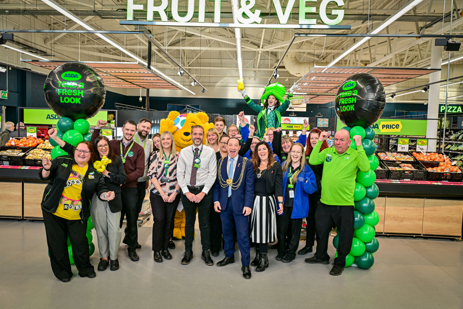 A group of ASDA staff stand in an ASDA store alongside Pudsey Bear. A sign up above state 'Fruit and Veg'