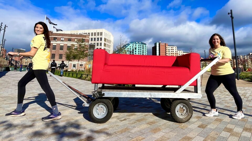 Two ladies in yellow t shirts pulling and pushing a large red sofa which is on top of a wheeled red trolley